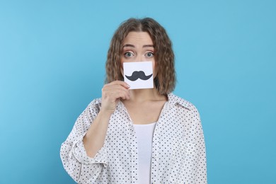 Photo of Woman holding paper with drawn mustache on light blue background