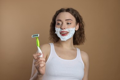 Photo of Happy woman with shaving foam on face holding razor against beige background