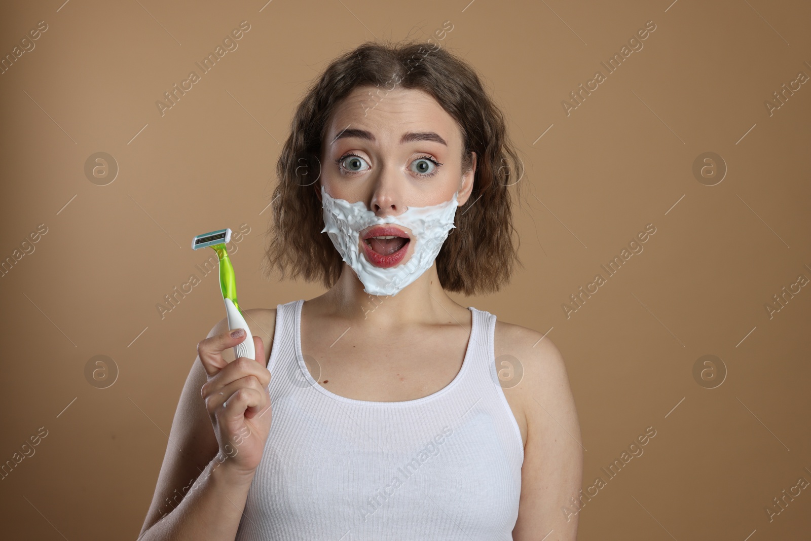 Photo of Emotional woman with shaving foam on face holding razor against beige background