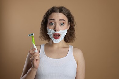 Photo of Emotional woman with shaving foam on face holding razor against beige background