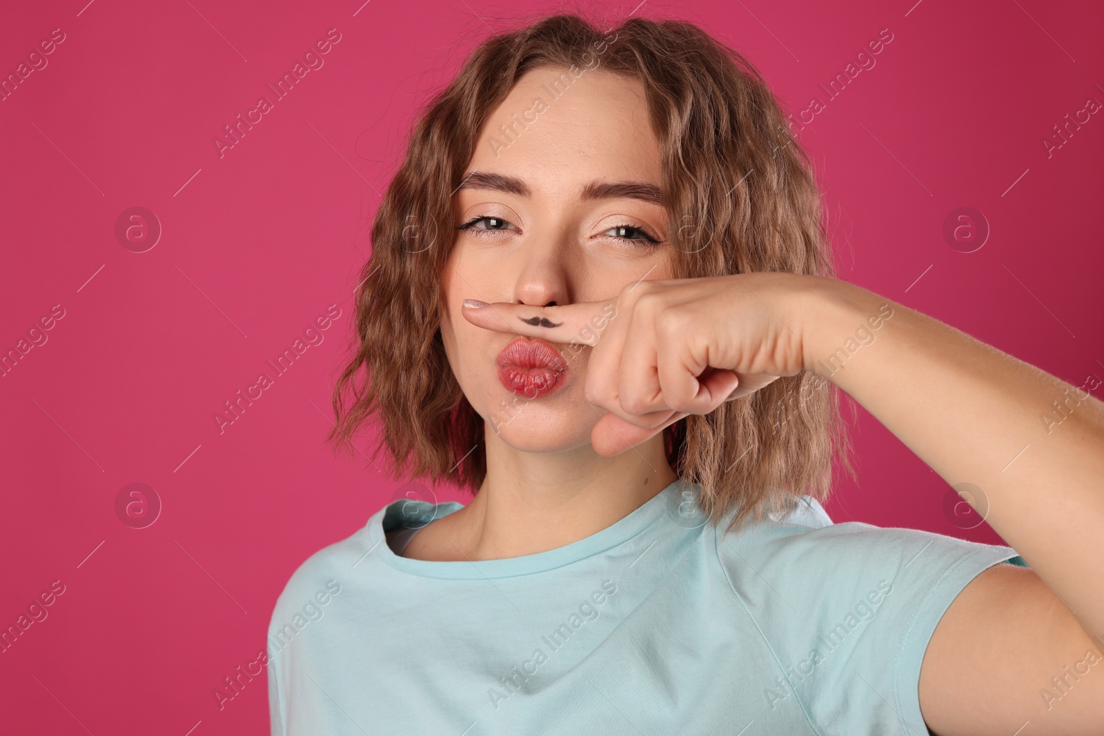 Photo of Funny woman holding finger with drawn mustache above lips on pink background