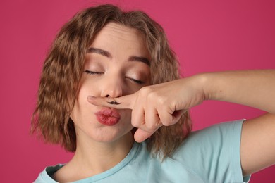 Photo of Funny woman holding finger with drawn mustache above lips on pink background