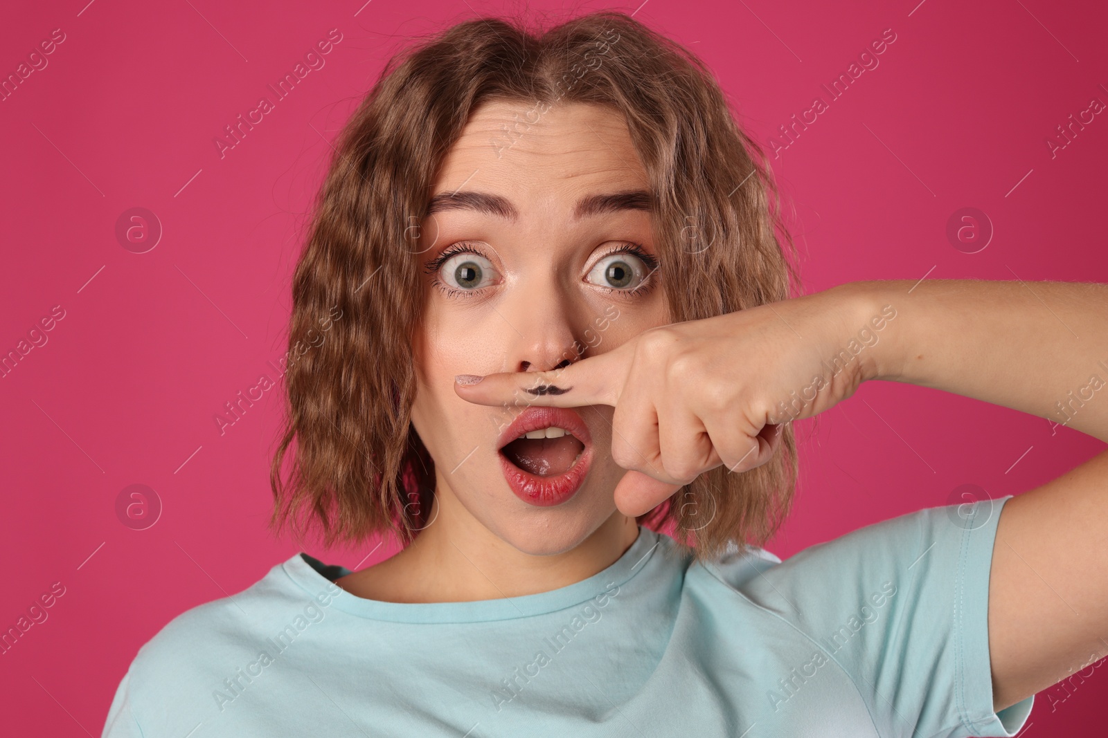 Photo of Emotional woman holding finger with drawn mustache above lips on pink background