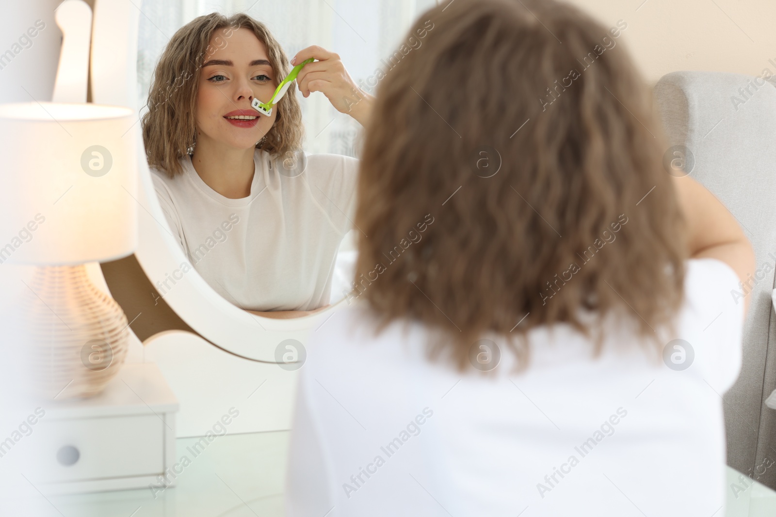 Photo of Happy woman shaving her mustache with razor near mirror at home