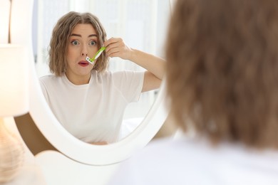 Photo of Emotional woman shaving her mustache with razor near mirror at home