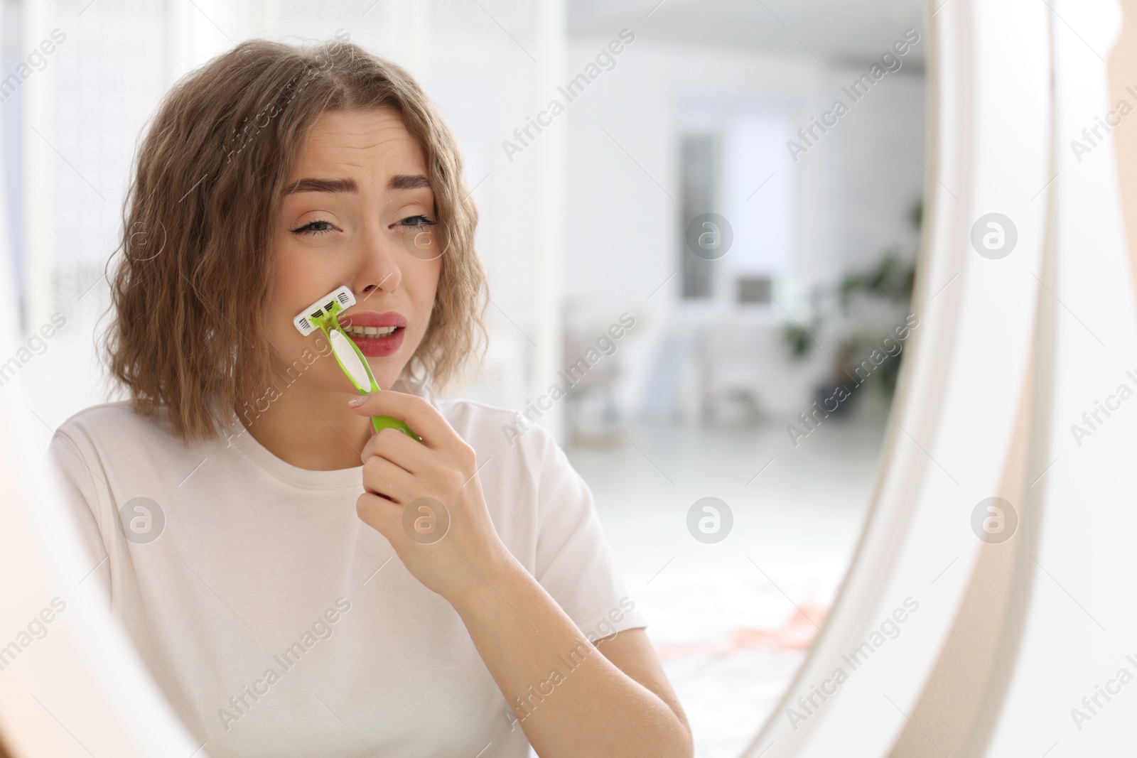 Photo of Beautiful woman shaving her mustache with razor near mirror at home