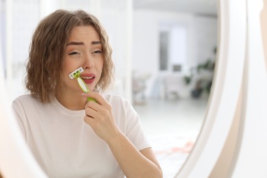 Photo of Beautiful woman shaving her mustache with razor near mirror at home