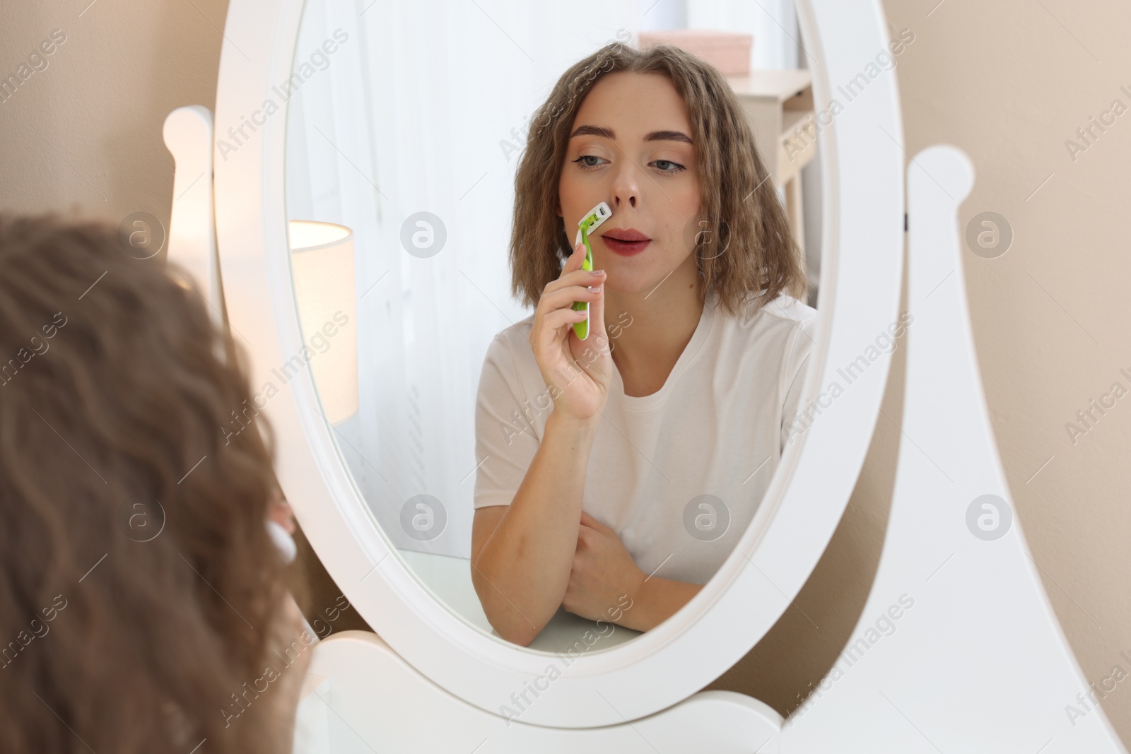 Photo of Beautiful woman shaving her mustache with razor near mirror at home