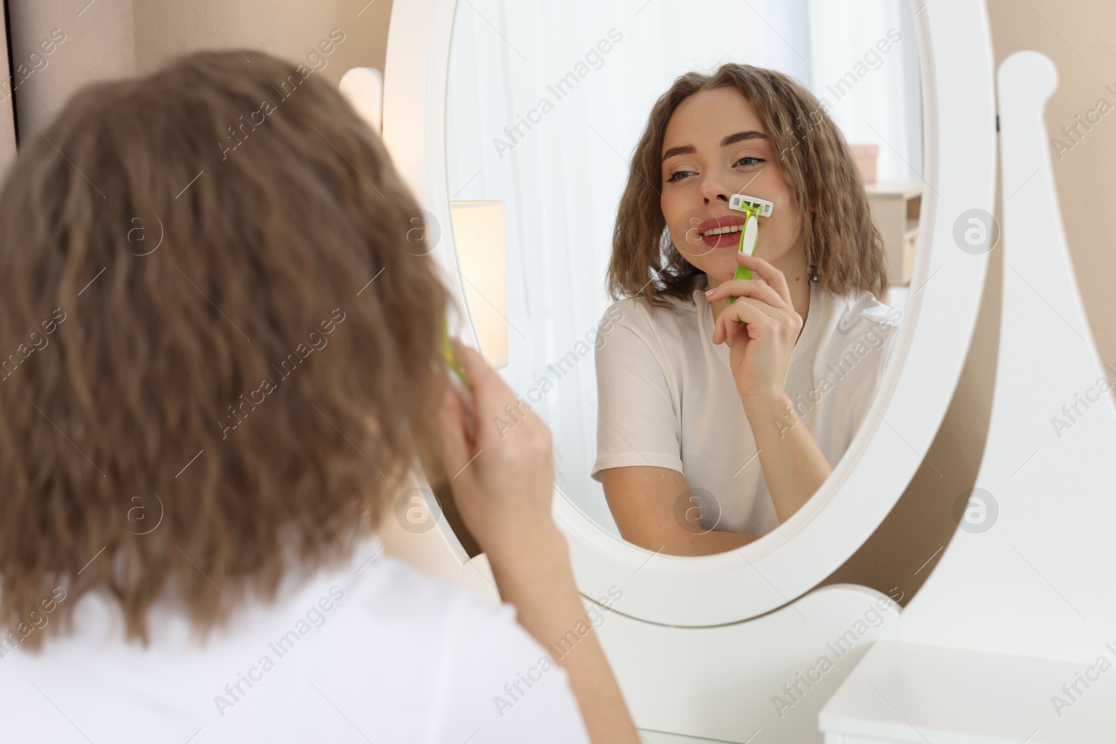 Photo of Happy woman shaving her mustache with razor near mirror at home