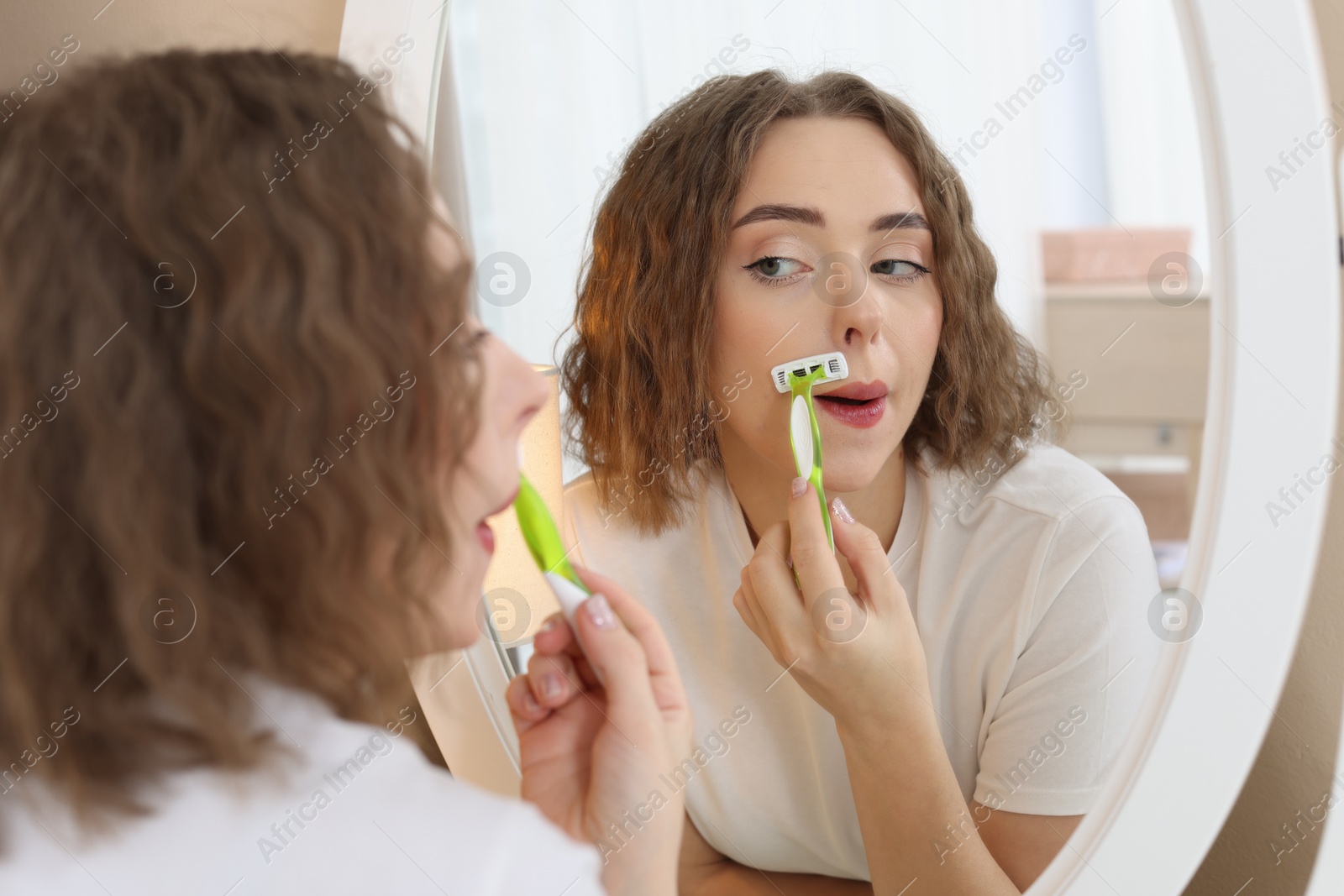 Photo of Beautiful woman shaving her mustache with razor near mirror at home