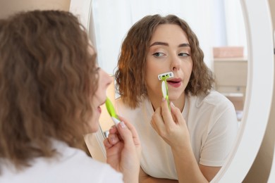 Photo of Beautiful woman shaving her mustache with razor near mirror at home