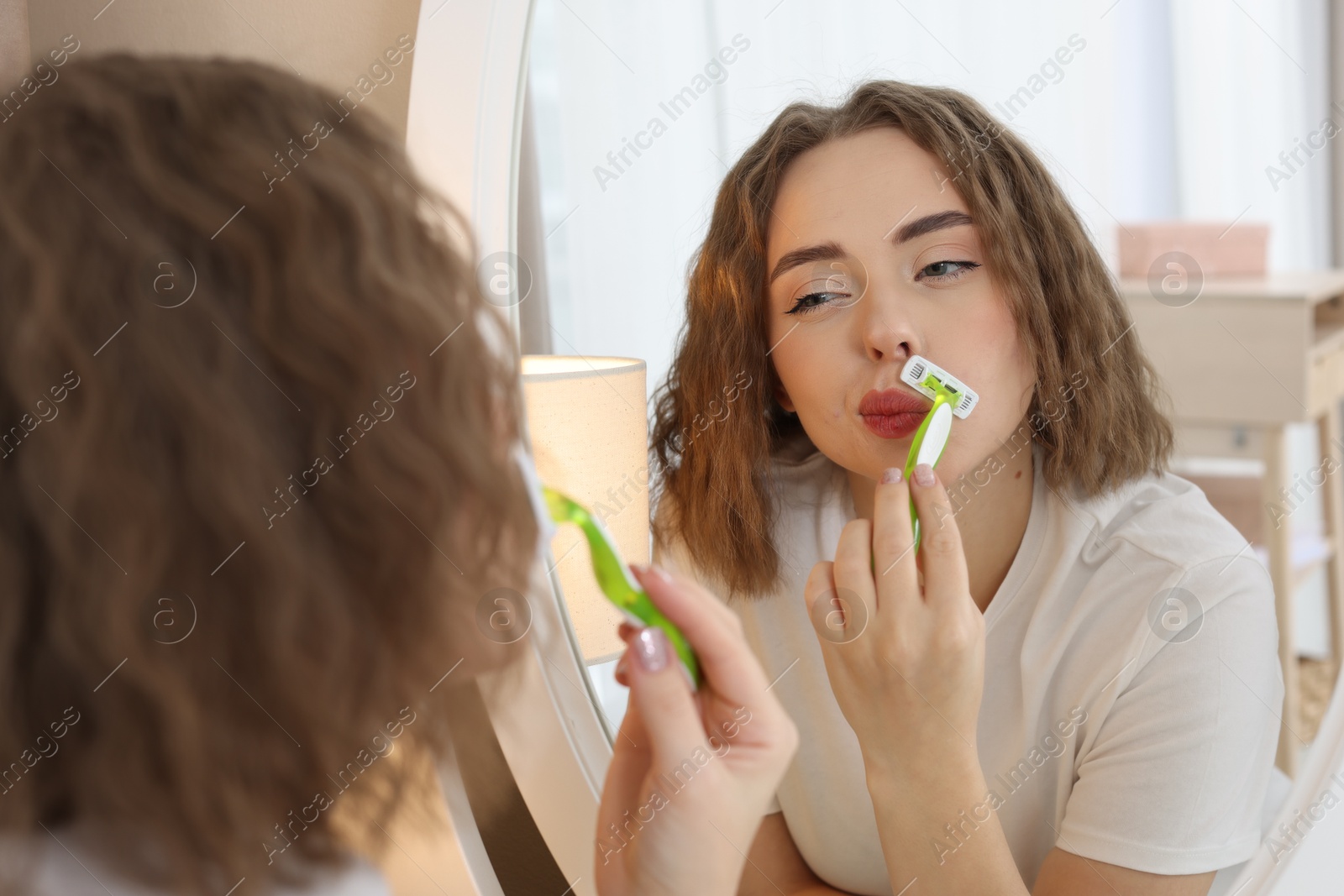 Photo of Beautiful woman shaving her mustache with razor near mirror at home