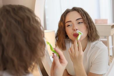 Photo of Beautiful woman shaving her mustache with razor near mirror at home