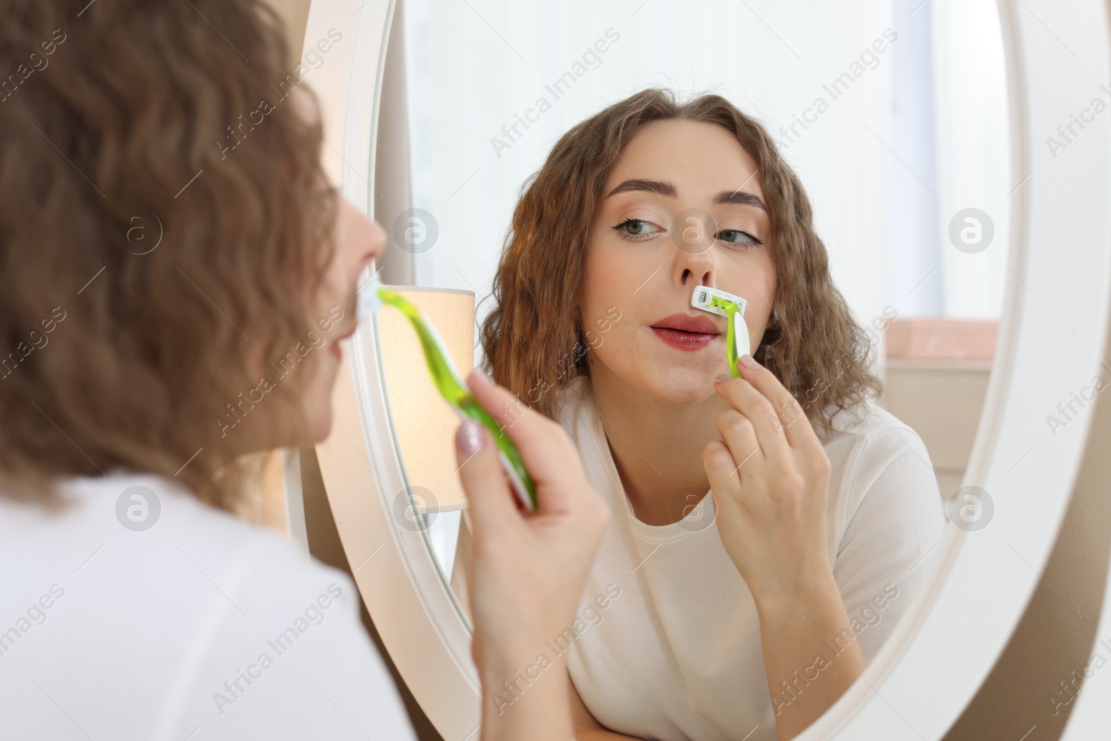 Photo of Beautiful woman shaving her mustache with razor near mirror at home