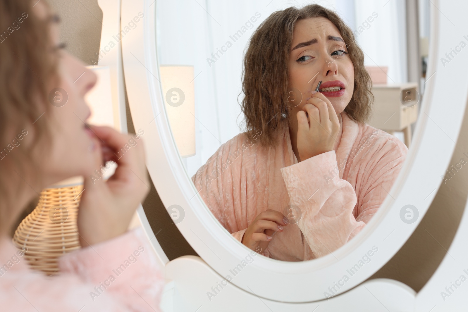 Photo of Beautiful woman plucking her mustache with tweezers near mirror at home