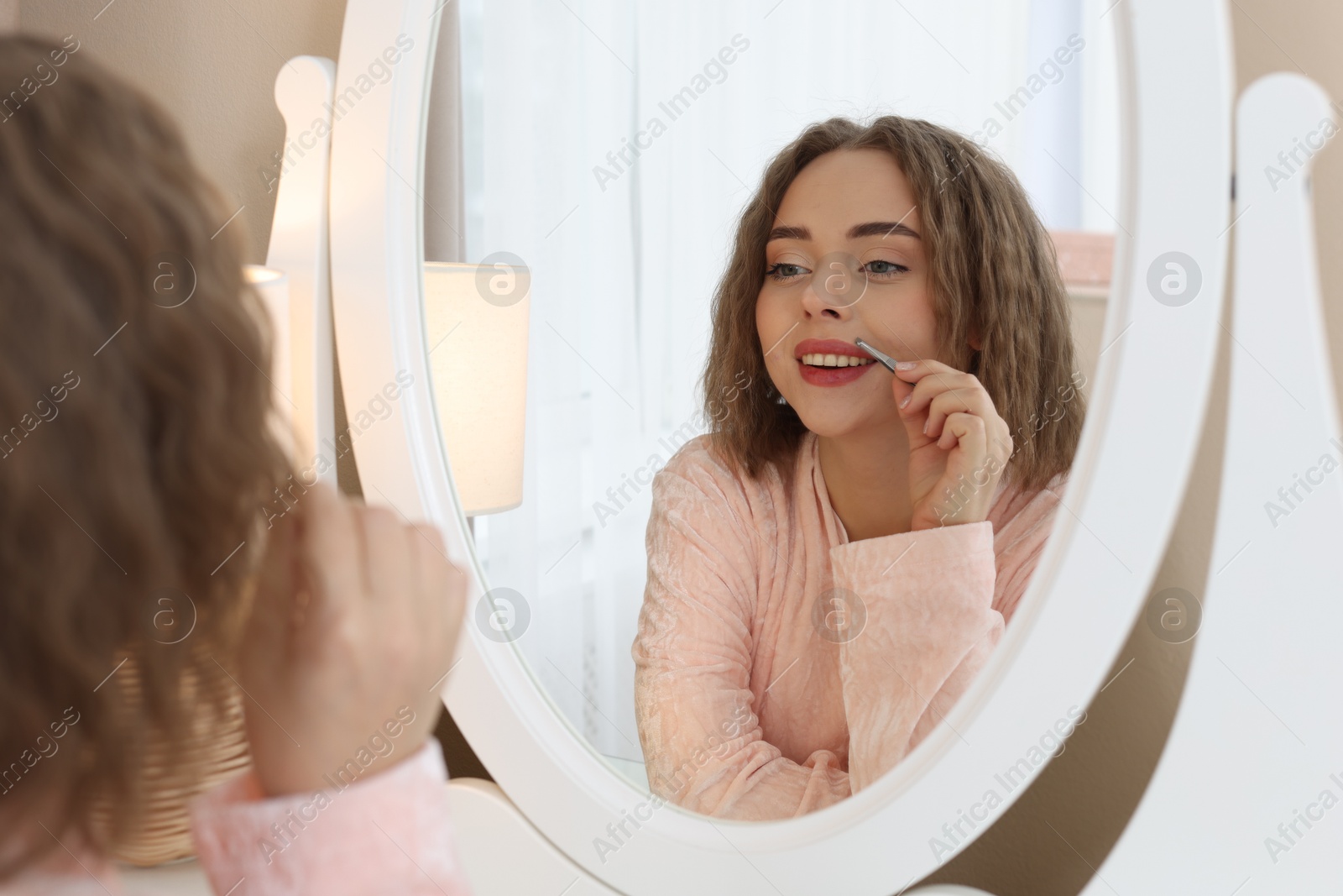 Photo of Happy woman plucking her mustache with tweezers near mirror at home