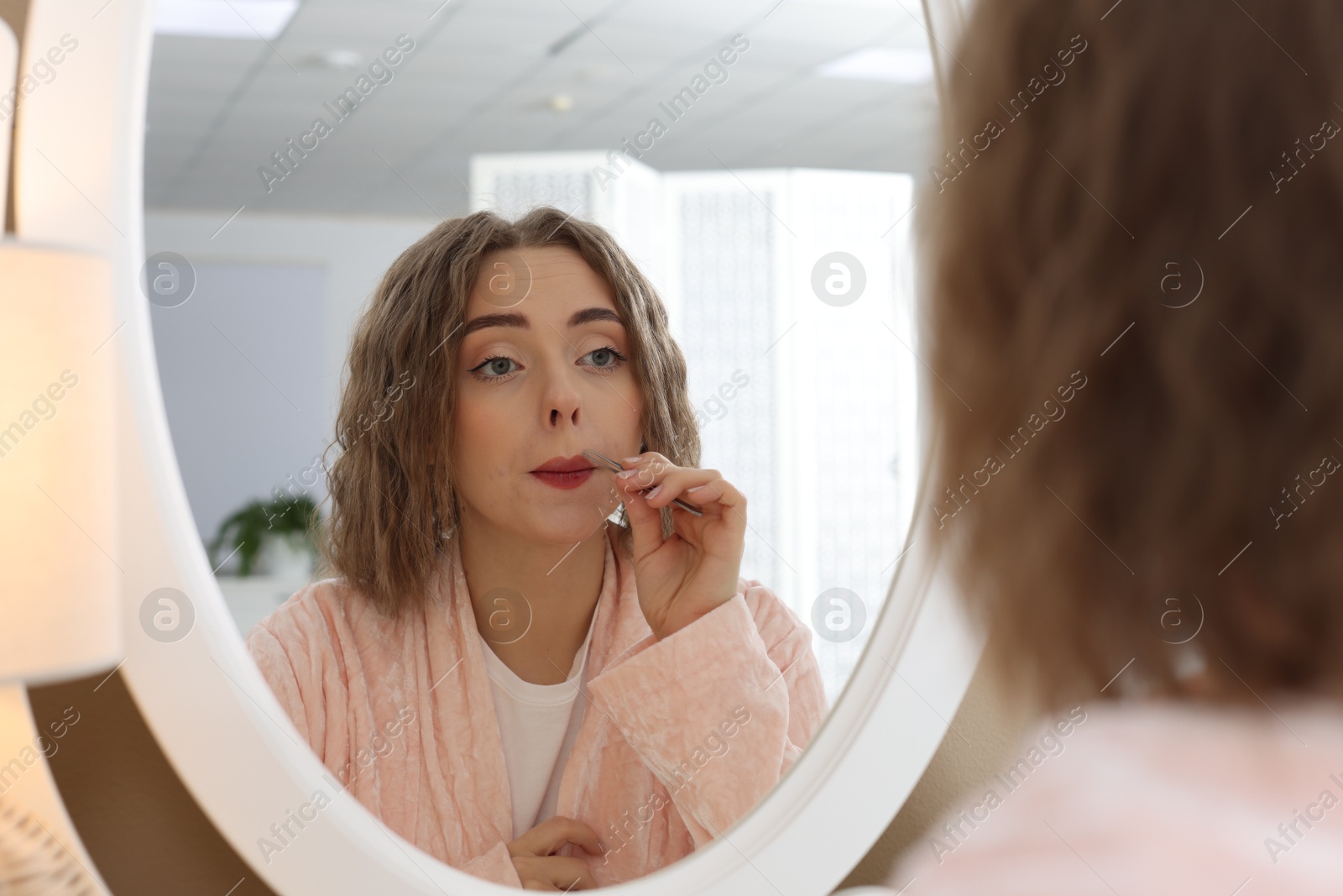 Photo of Beautiful woman plucking her mustache with tweezers near mirror at home