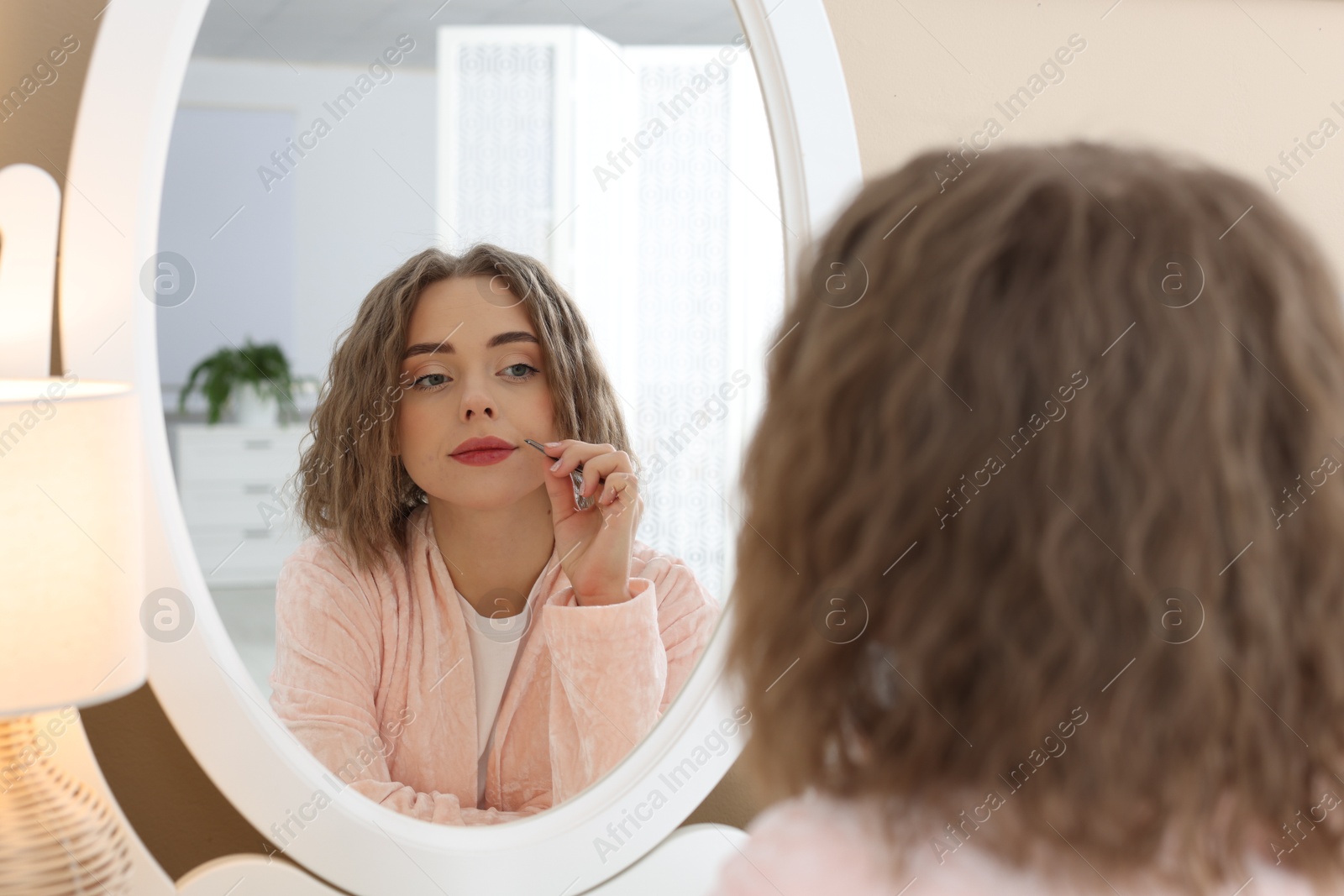 Photo of Beautiful woman plucking her mustache with tweezers near mirror at home