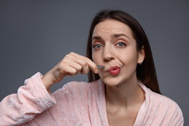 Photo of Beautiful woman plucking her mustache with tweezers on grey background