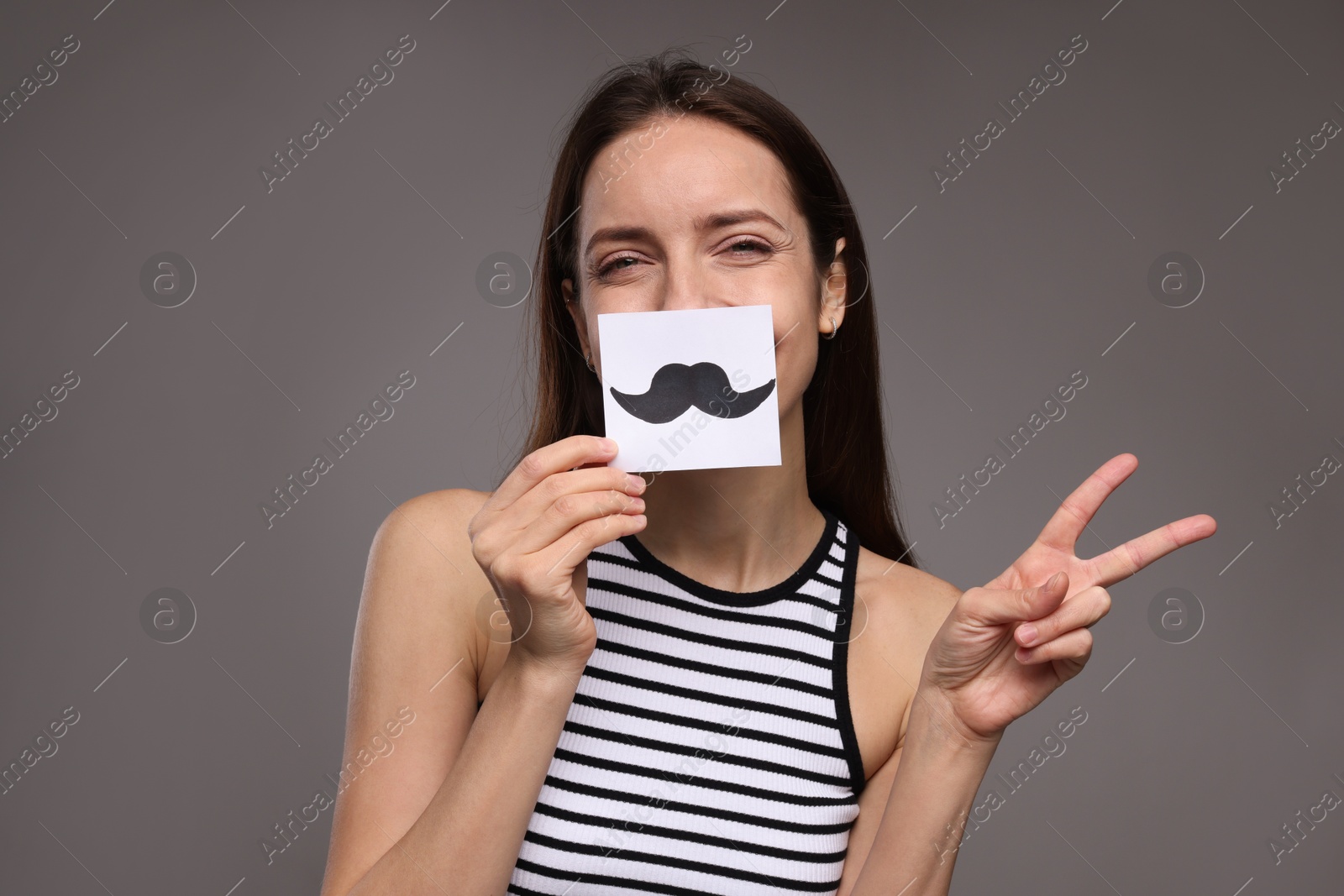 Photo of Woman holding paper with drawn mustache and showing peace sign on grey background