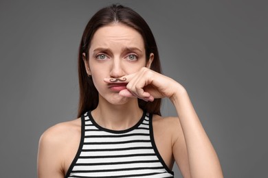 Photo of Sad woman holding finger with drawn mustache above lips on grey background