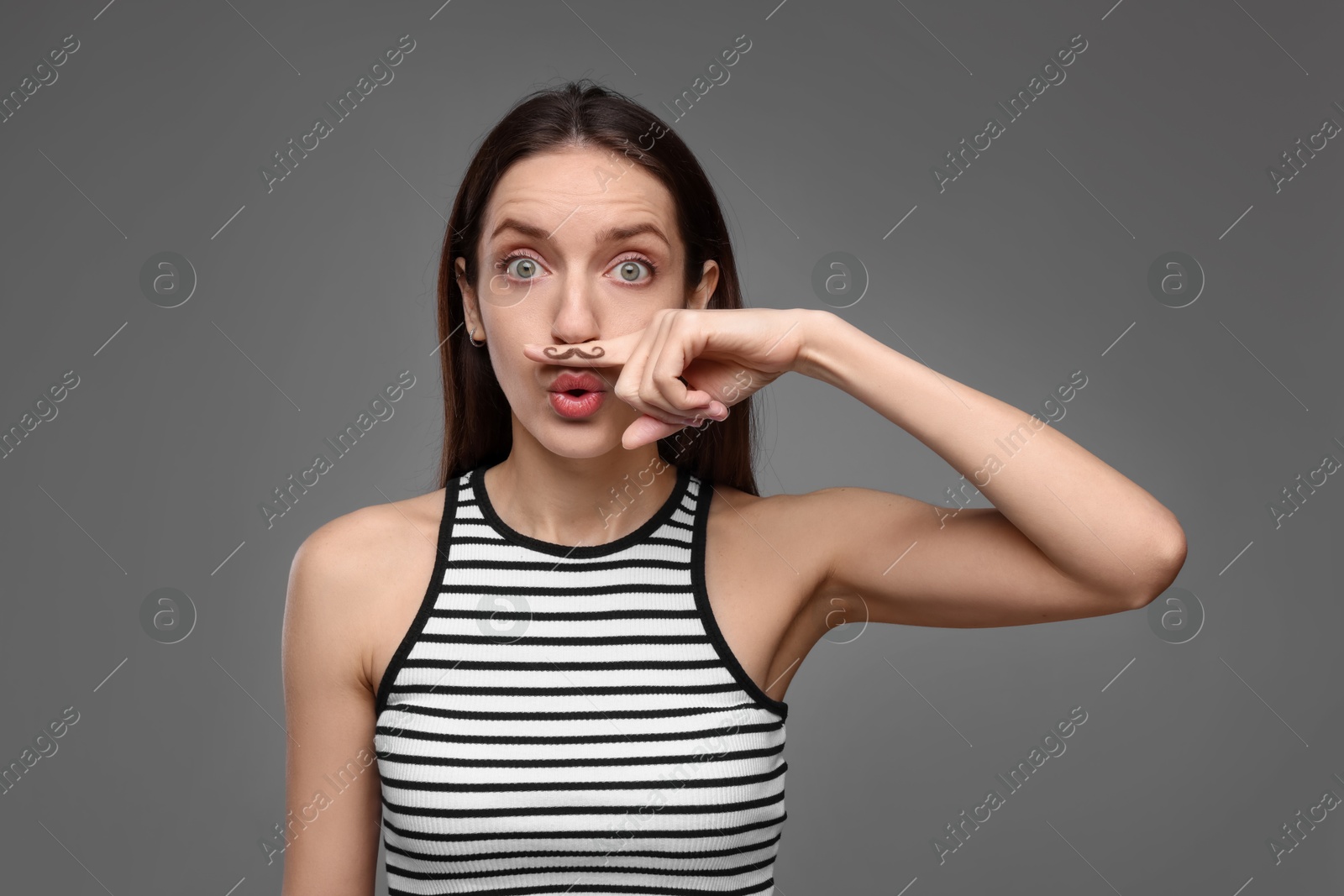 Photo of Funny woman holding finger with drawn mustache above lips on grey background