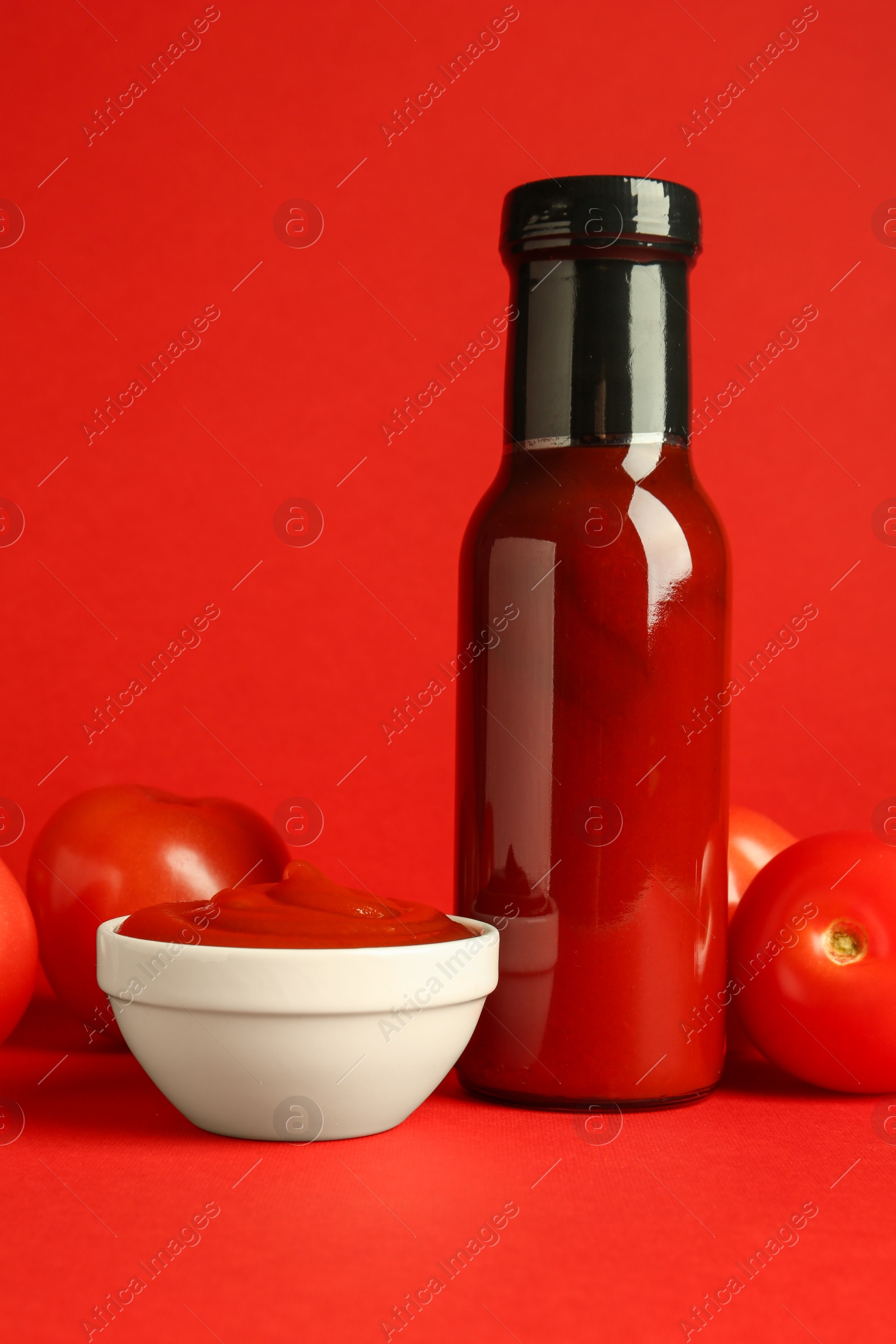 Photo of Ketchup in glass bottle, bowl and fresh tomatoes on red background