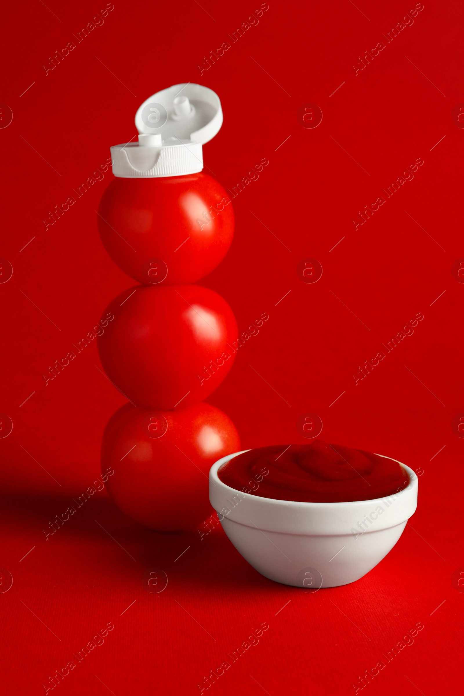 Photo of Stack of fresh tomatoes with plastic cap as bottle and ketchup in bowl on red background