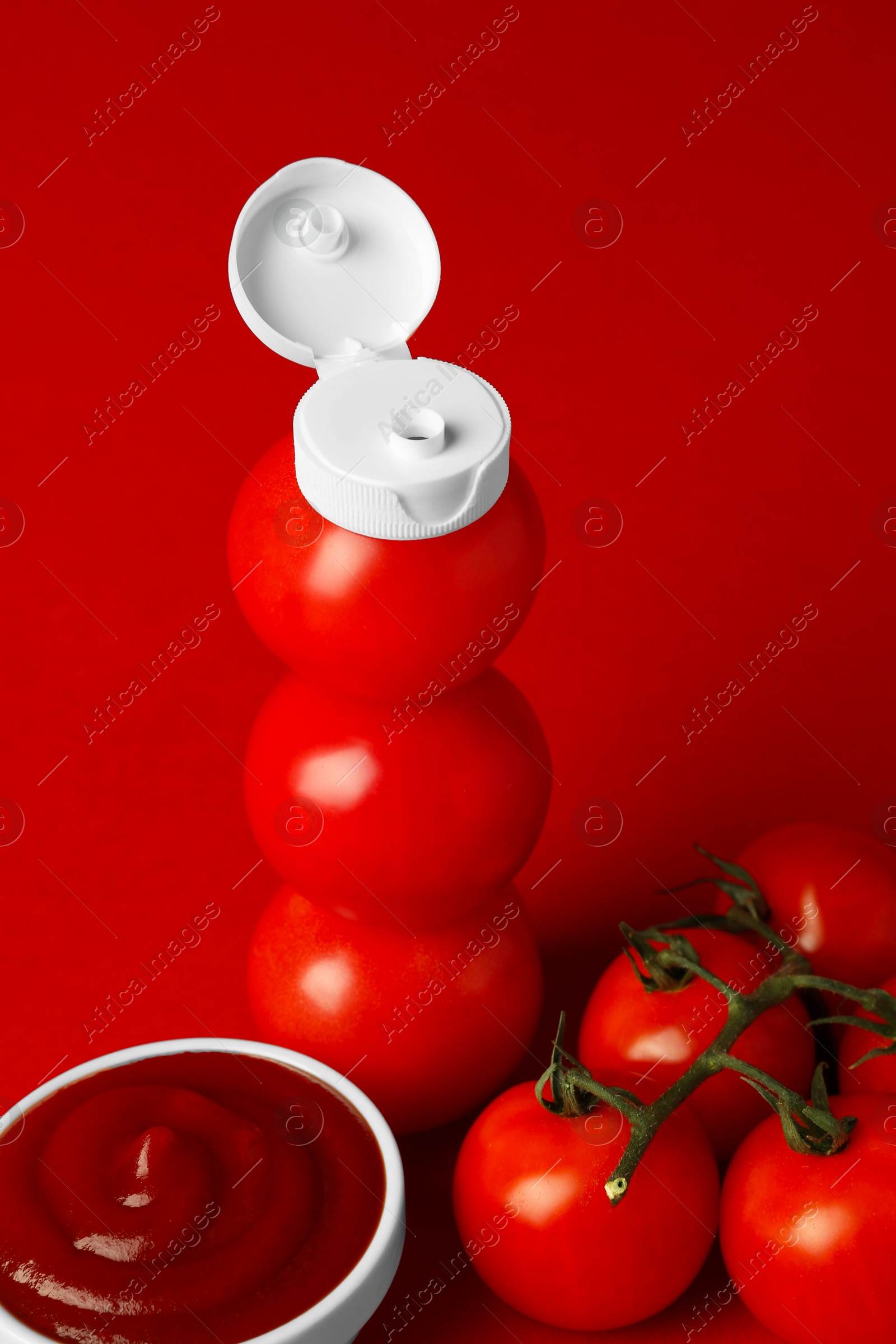 Photo of Stack of fresh tomatoes with plastic cap as bottle and ketchup in bowl on red background