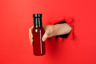 Photo of Woman holding bottle of ketchup through hole in torn red paper, closeup