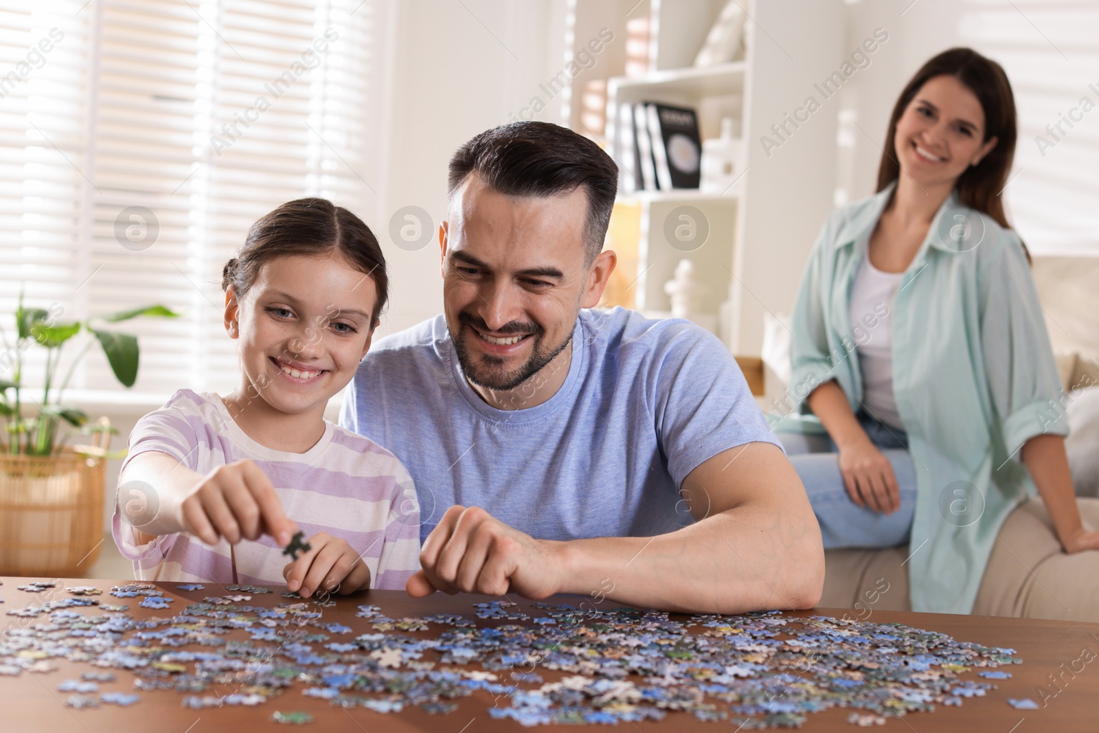 Photo of Happy father and his daughter solving puzzle together at wooden table while mother sitting on couch indoors