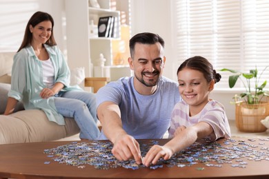 Photo of Happy father and his daughter solving puzzle together at wooden table while mother sitting on couch indoors