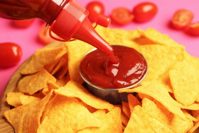 Photo of Pouring tasty ketchup in bowl with nachos on pink background, closeup