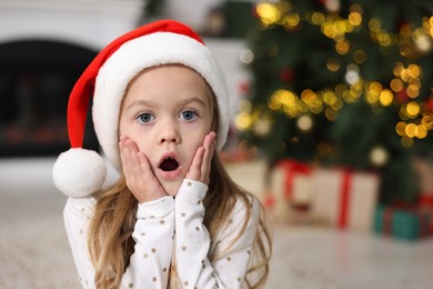 Portrait of emotional little girl in Santa hat against Christmas lights