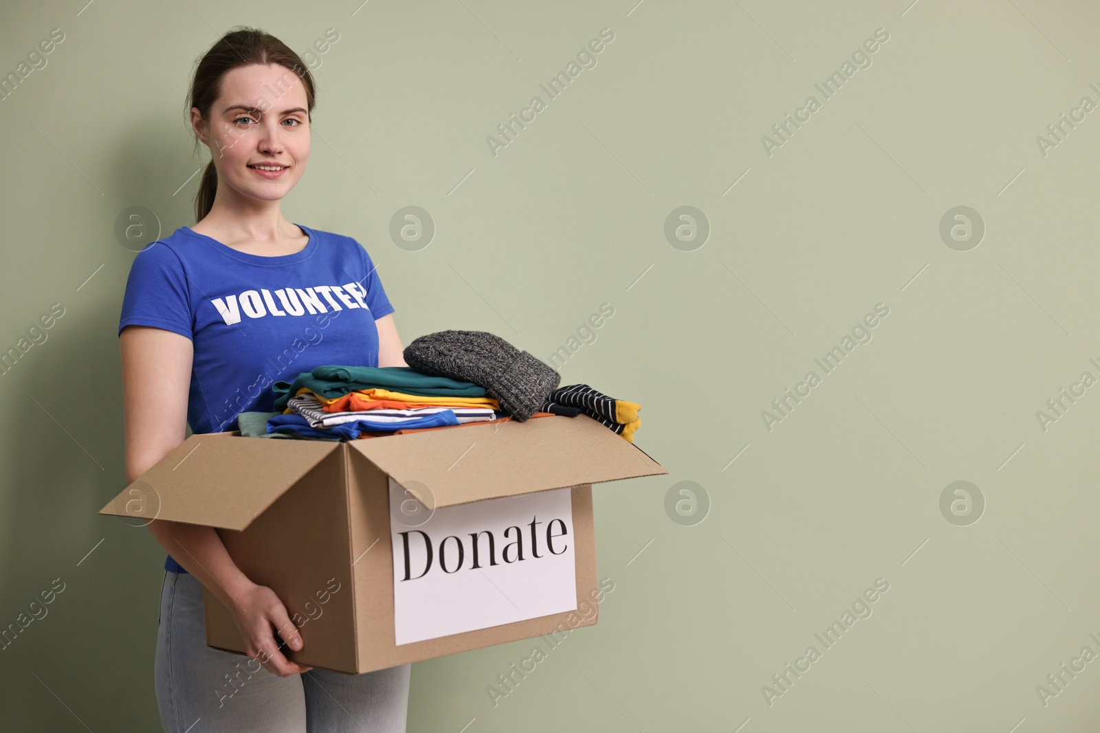 Photo of Woman holding donation box with clothes on pale olive background, space for text