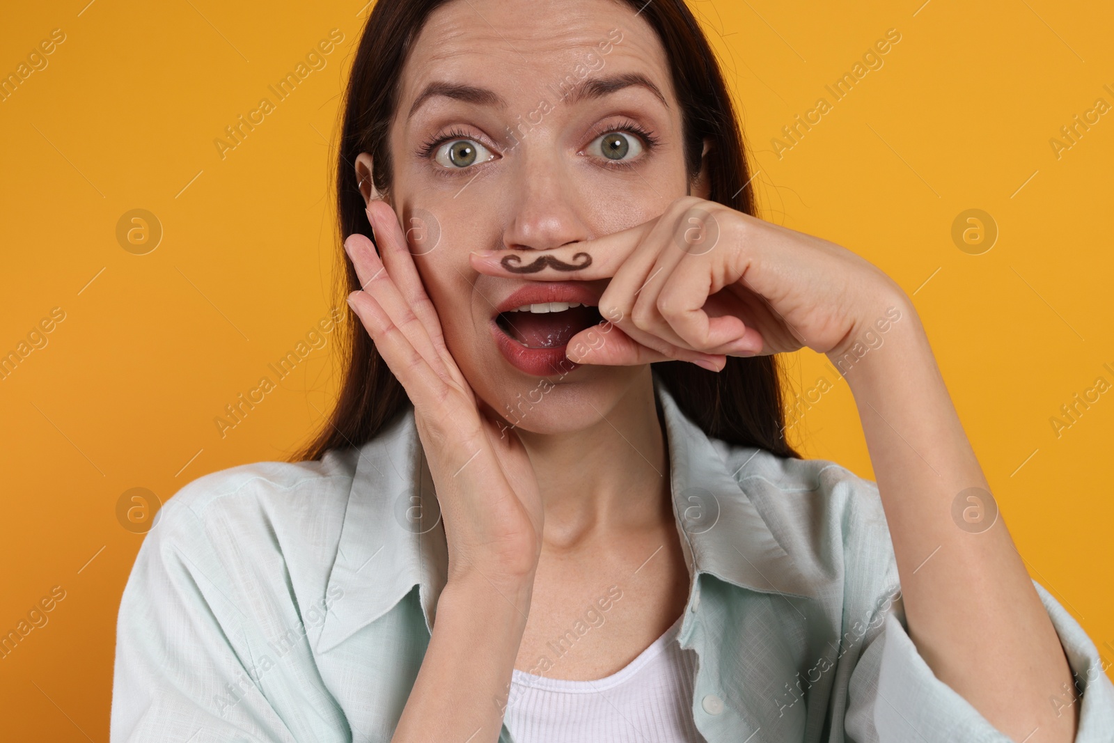 Photo of Emotional woman holding finger with drawn mustache above lips on orange background