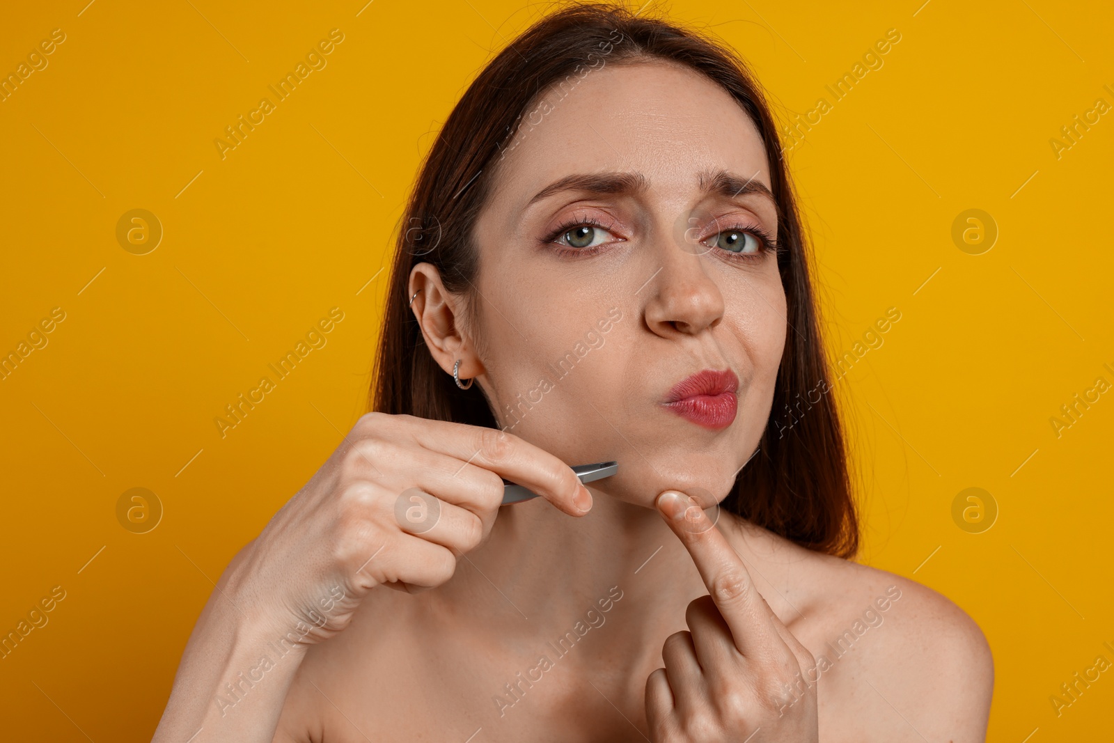 Photo of Beautiful woman plucking her facial hair with tweezers on orange background