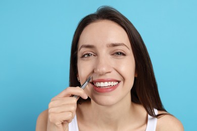 Happy woman plucking her mustache with tweezers on light blue background