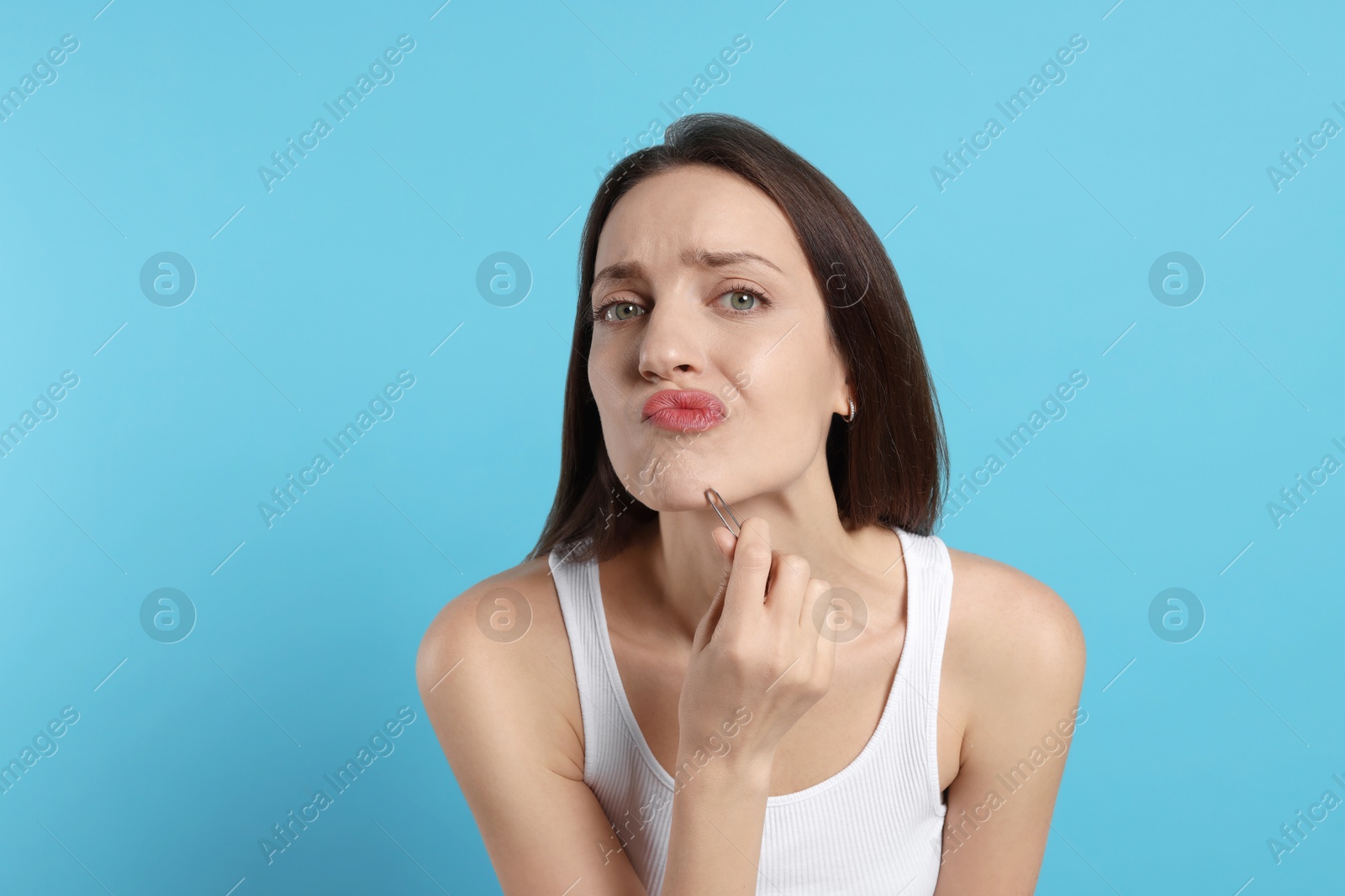 Photo of Woman plucking her facial hair with tweezers on light blue background