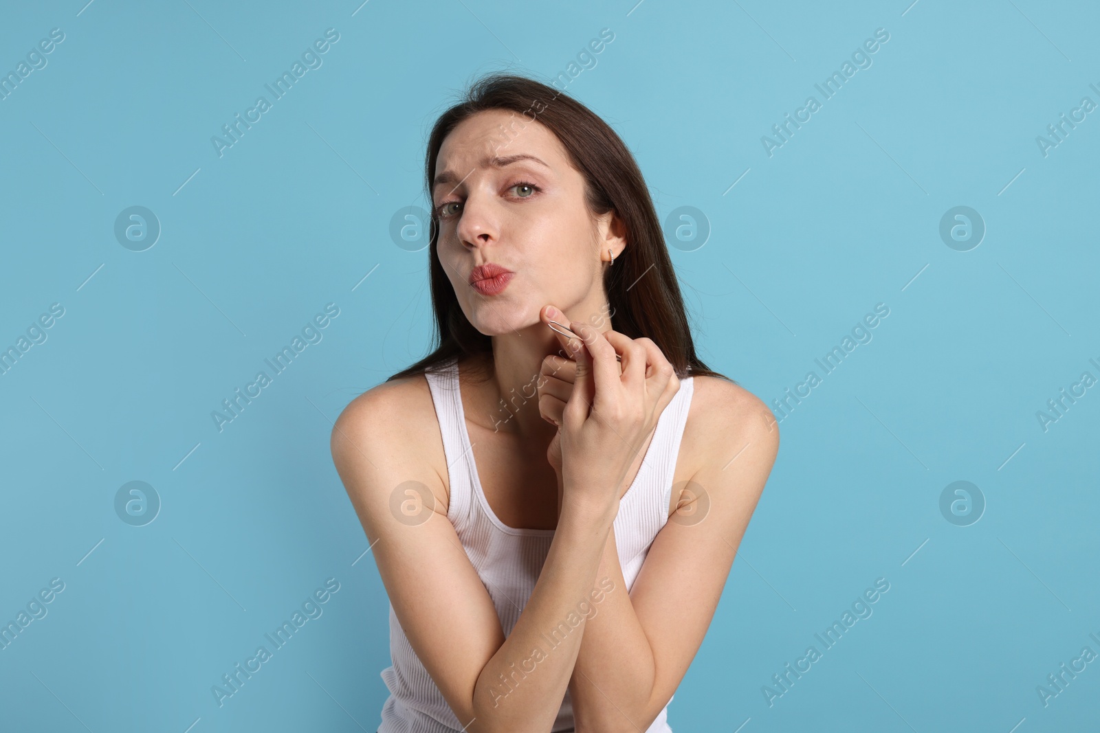 Photo of Woman plucking her facial hair with tweezers on light blue background