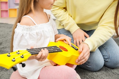 Photo of Woman teaching little girl to play ukulele at home, closeup