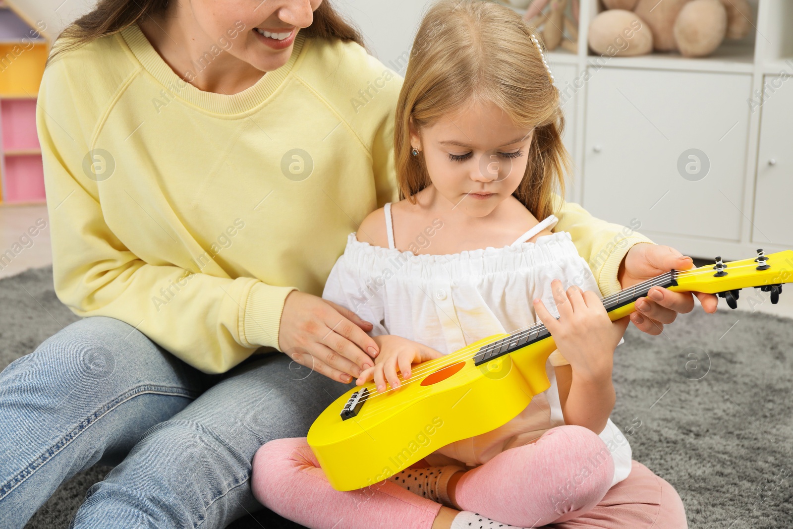 Photo of Young woman teaching little girl to play ukulele at home, closeup