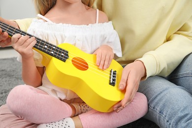 Photo of Young woman teaching little girl to play ukulele at home, closeup