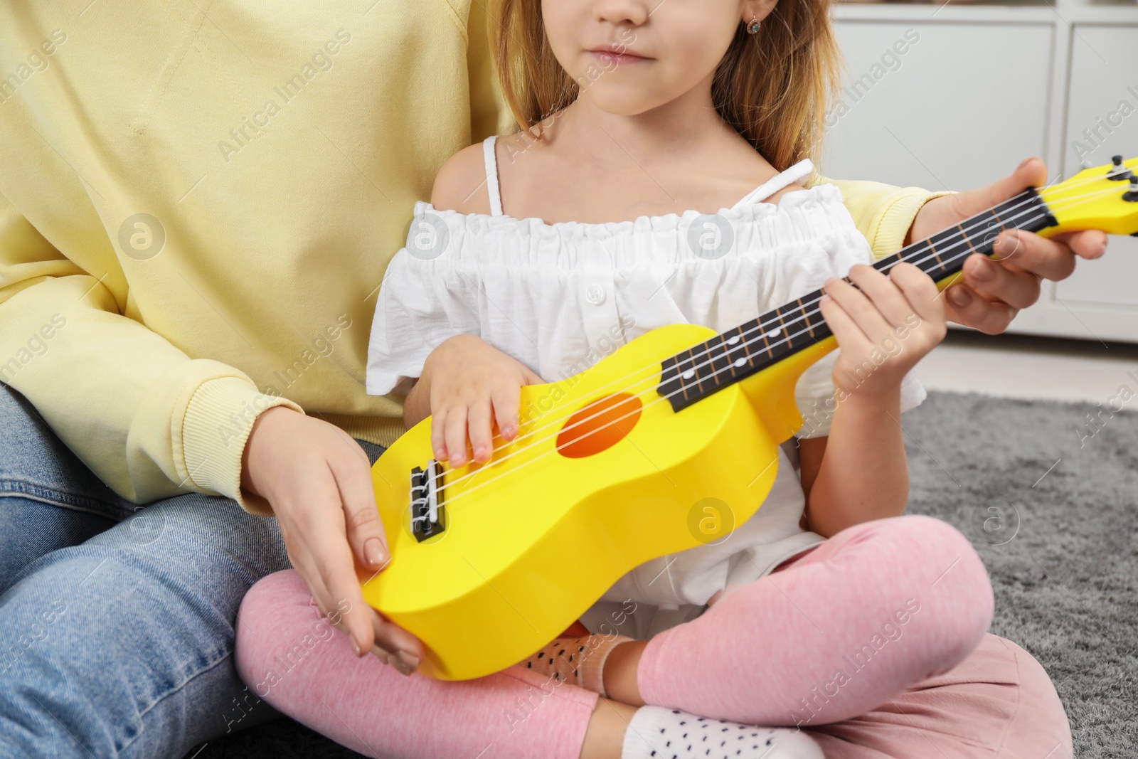 Photo of Young woman teaching little girl to play ukulele at home, closeup