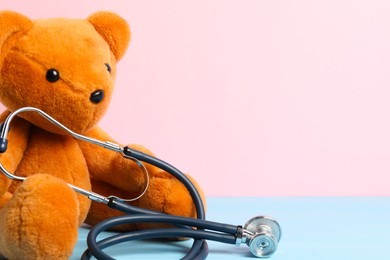 Photo of Pediatrics concept. Teddy bear with stethoscope on light blue wooden table against pink background, space for text