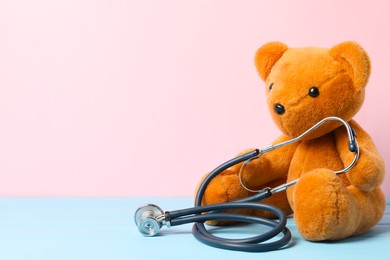 Photo of Pediatrics concept. Teddy bear with stethoscope on light blue wooden table against pink background, space for text