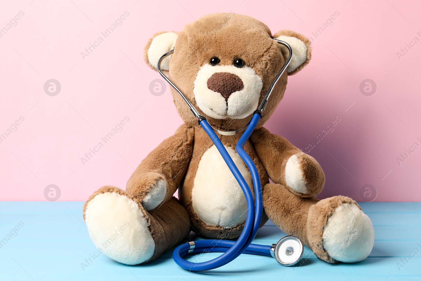 Photo of Pediatrics concept. Teddy bear with stethoscope on light blue wooden table against pink background