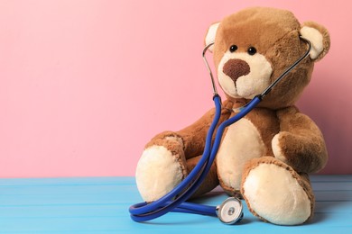 Photo of Pediatrics concept. Teddy bear with stethoscope on light blue wooden table against pink background, space for text