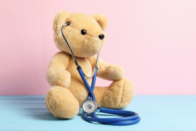 Photo of Pediatrics concept. Teddy bear with stethoscope on light blue wooden table against pink background, space for text