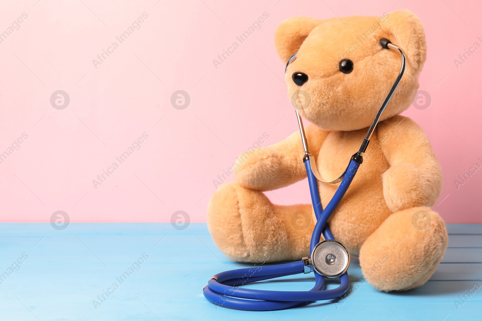 Photo of Pediatrics concept. Teddy bear with stethoscope on light blue wooden table against pink background, space for text